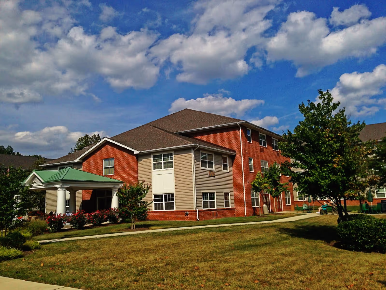 Multi-story brick and siding senior living building with a covered entrance, lawn, trees, and a partly cloudy blue sky.