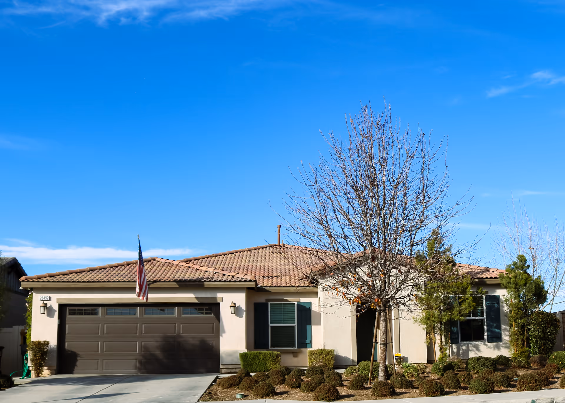 A single-story beige house with a two-car garage, flag, and landscaped front yard under blue sky.