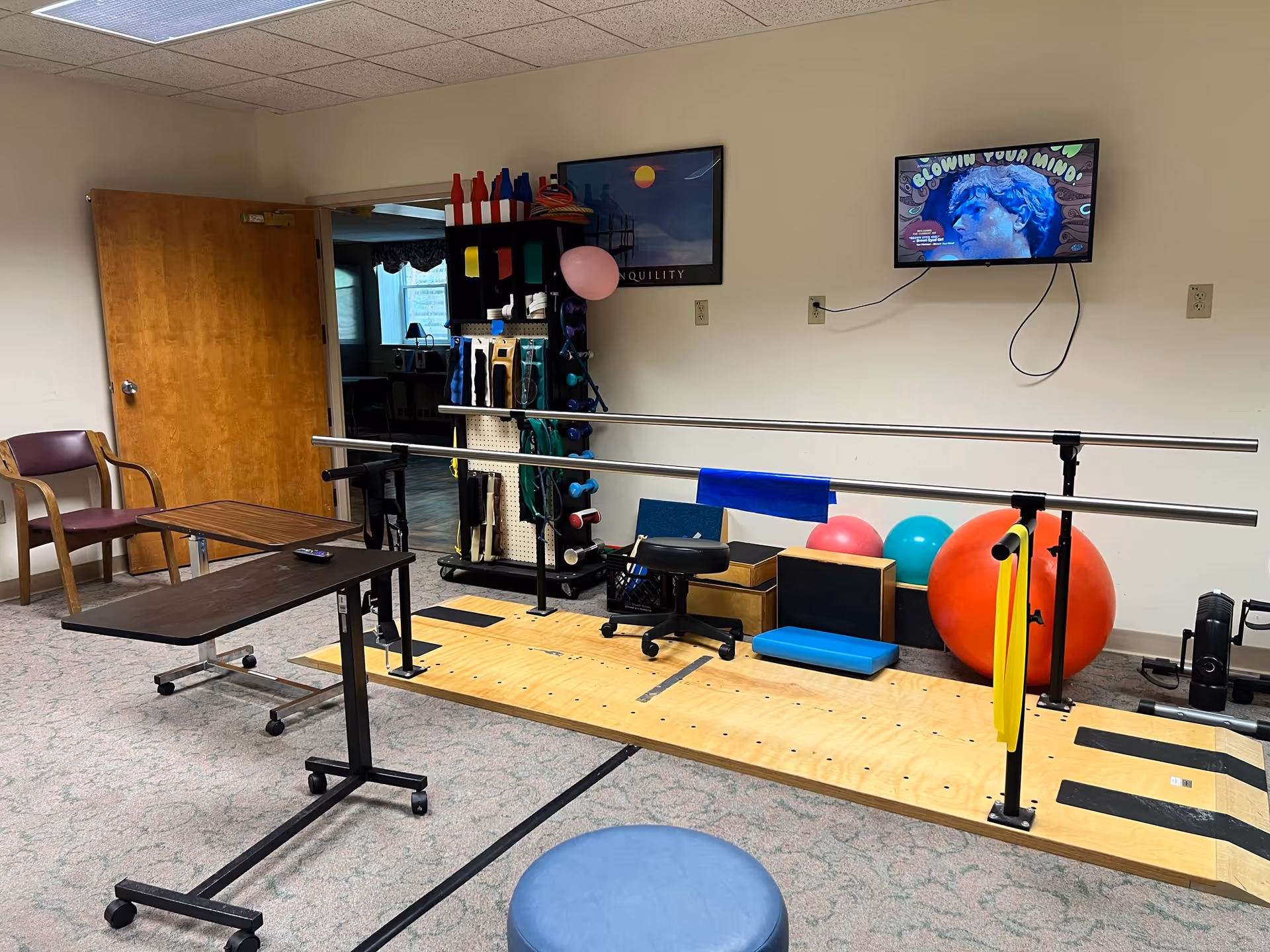 A therapy or rehabilitation room with parallel bars for walking exercises, various exercise balls, therapy equipment on a pegboard, a wooden platform, a chair, and two adjustable overbed tables. A TV is mounted on the wall displaying an image with the text 'BLOWIN YOUR MIND'.