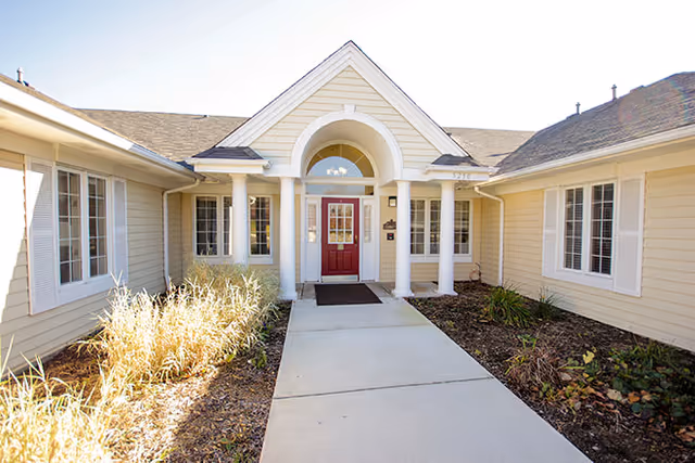 Front entrance of a single-story building with beige siding, white trim, and a red door under a small portico supported by four white columns. There are windows on either side of the door and a concrete walkway leading up to the entrance, with landscaping on both sides.