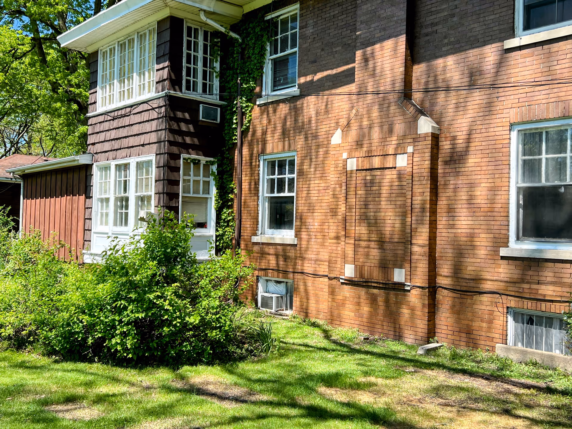 Exterior view of a senior living facility building with brown brick and wooden siding. The building has multiple white-framed windows, some with air conditioning units installed. There is greenery and bushes in front of the building, and sunlight casts shadows on the walls and grass.