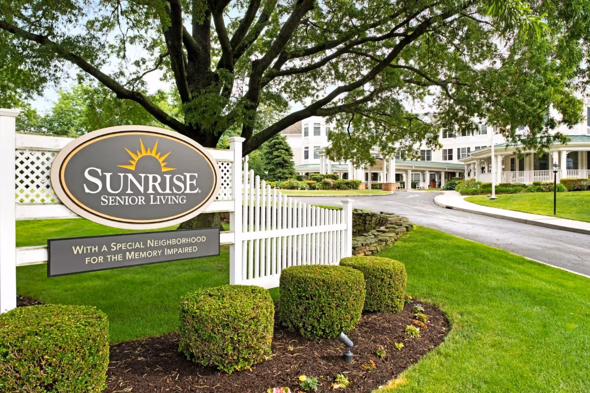 Entrance sign for Sunrise Senior Living on a landscaped lawn with the facility building and driveway in the background.