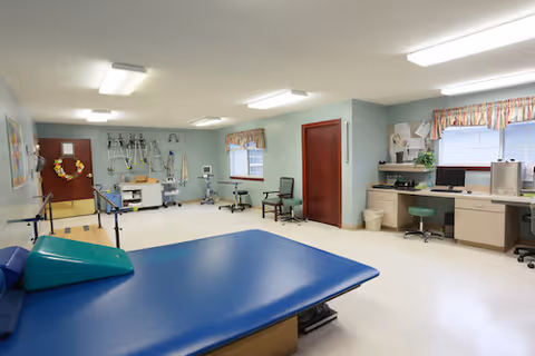 A spacious physical therapy or rehabilitation room with a large blue therapy table in the foreground, exercise equipment along the back wall, two windows with colorful valances, a desk with a computer and office chair on the right side, and light blue walls with white flooring.
