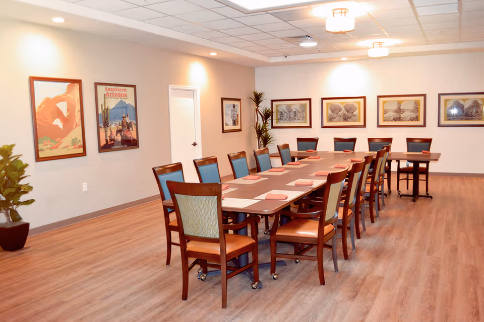 A spacious dining room with a long rectangular table surrounded by wooden chairs with blue and beige upholstery. The table is set with placemats and folded napkins. The room has light-colored walls adorned with framed artwork and posters, and there are potted plants in the corners. The floor is wood, and the ceiling has recessed lighting and round light fixtures.
