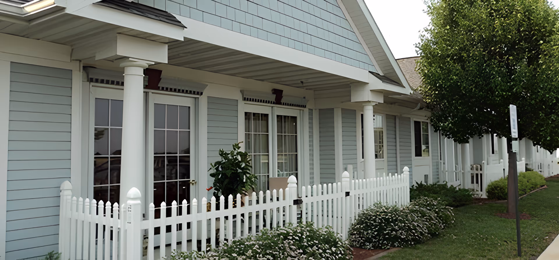 Exterior front of a pale-blue building with a covered porch, white picket fence, columns, and landscaped shrubs.
