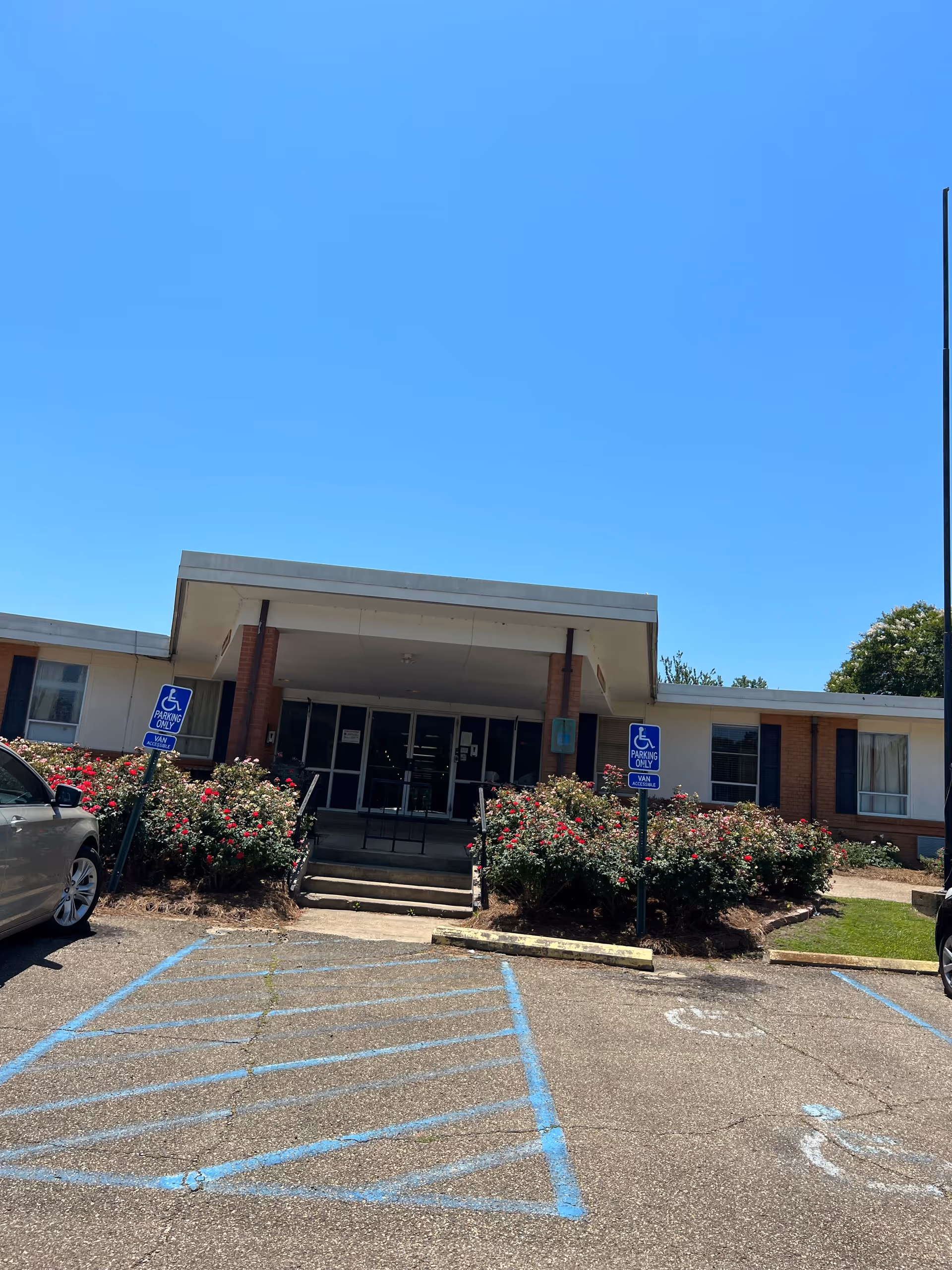 Front entrance of a single-story brick retirement facility with wheelchair parking spaces, steps up to glass doors, and rose beds under a clear blue sky.