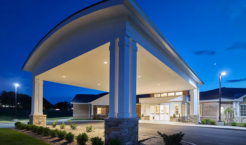 Lit covered entrance porte-cochere of a senior living facility at dusk with landscaped grounds and front doors visible.