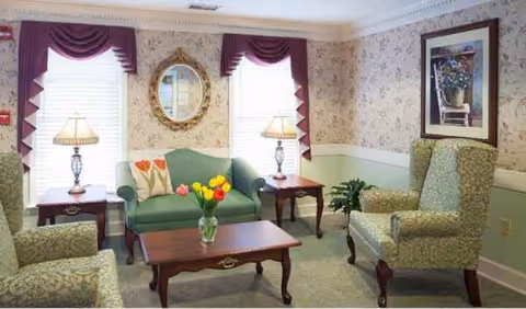 Cozy senior living lounge with green sofa, armchairs, coffee table, lamps, tulips, and curtains.