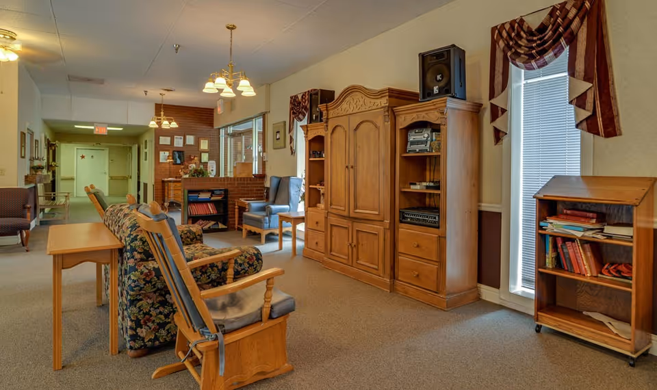 A cozy nursing center common area with wooden furniture including a floral-patterned sofa, cushioned chairs, a wooden entertainment center with audio equipment, and a small bookshelf. The room has beige walls, carpeted floor, two windows with valances, and ceiling lights. A hallway with an exit sign is visible in the background.
