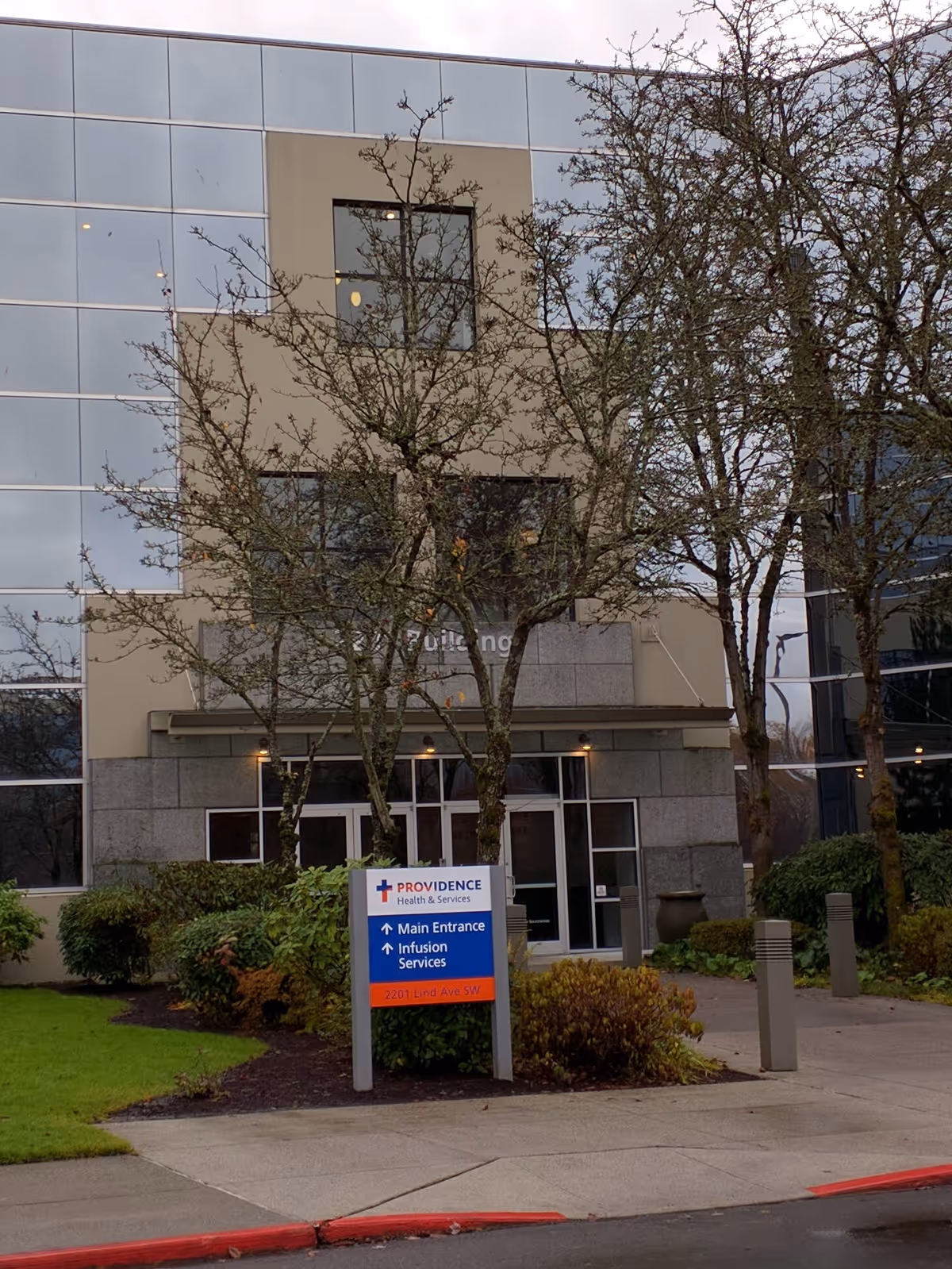 Modern glass-front building exterior with entrance sign and leafless trees in front.