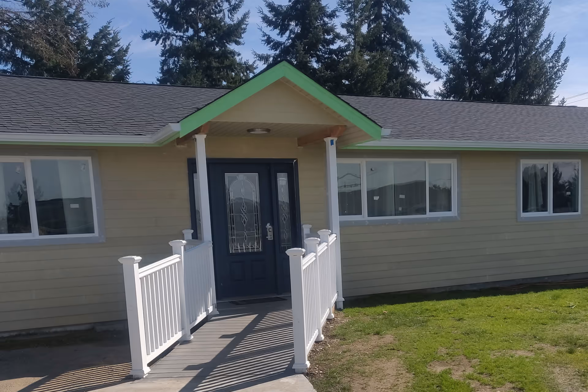 Exterior view of a single-story building with beige siding and a dark blue front door with decorative glass panels. The entrance has a small covered porch with a green-trimmed roof and white railings along a concrete ramp leading to the door. There are several windows on the front of the building and tall evergreen trees in the background under a clear sky.
