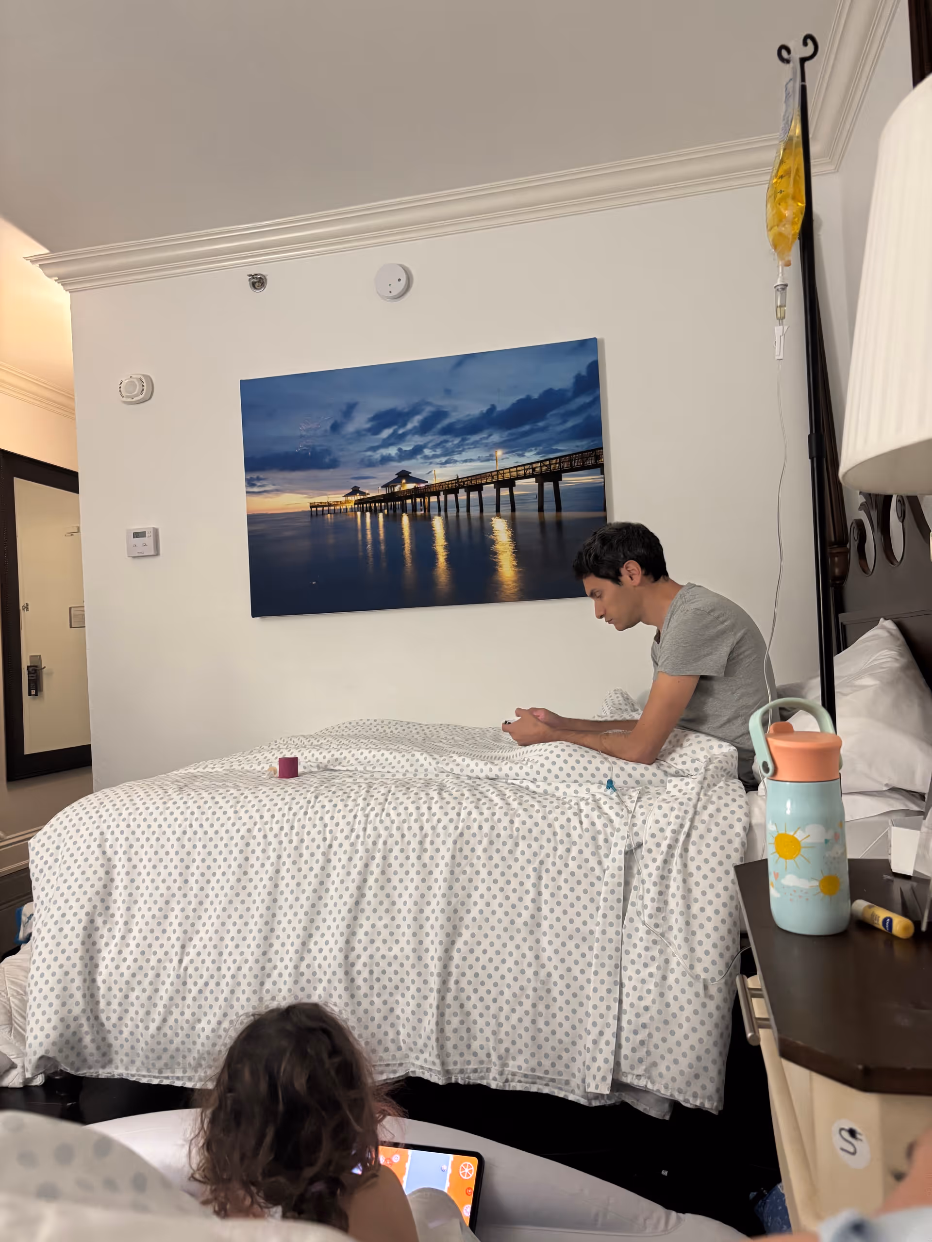 Two people sit on a polka-dotted bed in a bright hospital-style bedroom with IV drip.