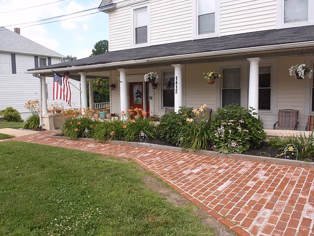 Hampton Meadows Assisted Living exterior featuring a covered porch, columns, flag, and landscaped flowers.