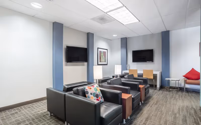 Modern senior living lounge with black leather chairs, TVs, lamps, and blue accent columns.