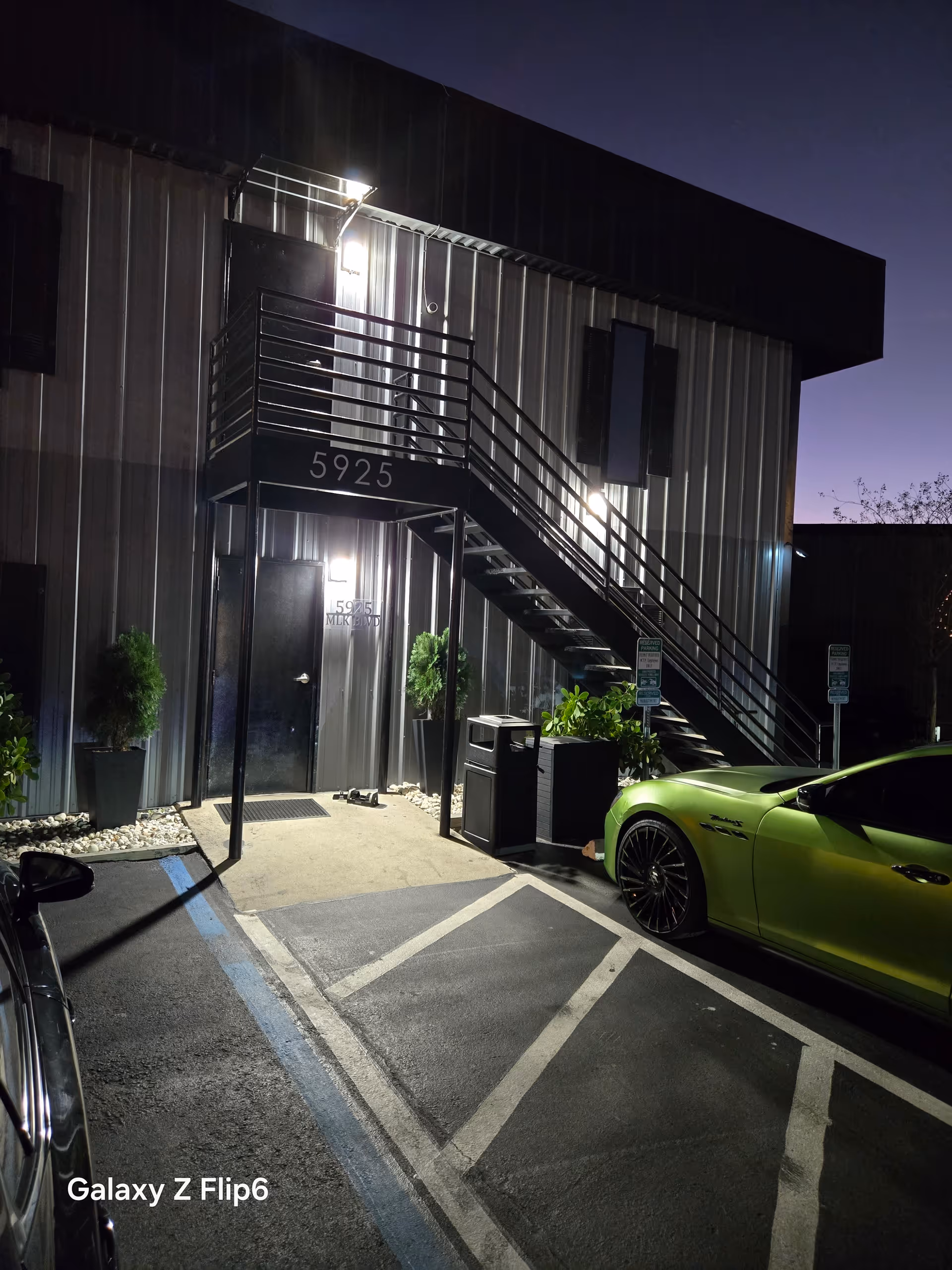 Nighttime exterior of a metal building with external stairs, parking lot, and a lime-green car.