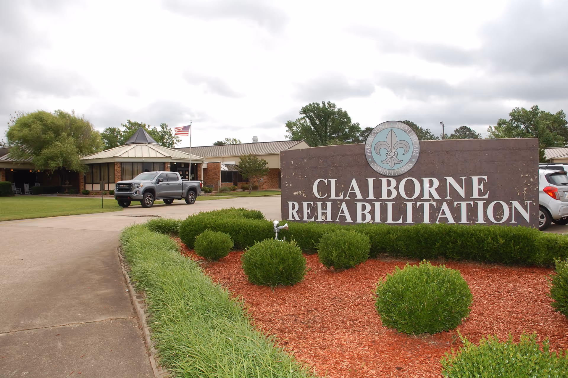Exterior view of Claiborne Rehabilitation facility with a large sign in the foreground surrounded by neatly trimmed bushes and red mulch. A gray pickup truck is parked on the driveway leading to the building, which has a metal roof and an American flag flying on a flagpole. The sky is cloudy.