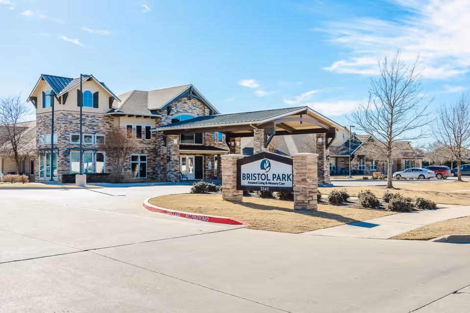 Exterior view of Bristol Park at Eagle Mountain Assisted Living & Memory Care facility with a stone and brick building, a covered entrance, a sign displaying the facility name and address, a parking area with cars, and leafless trees on a clear day.