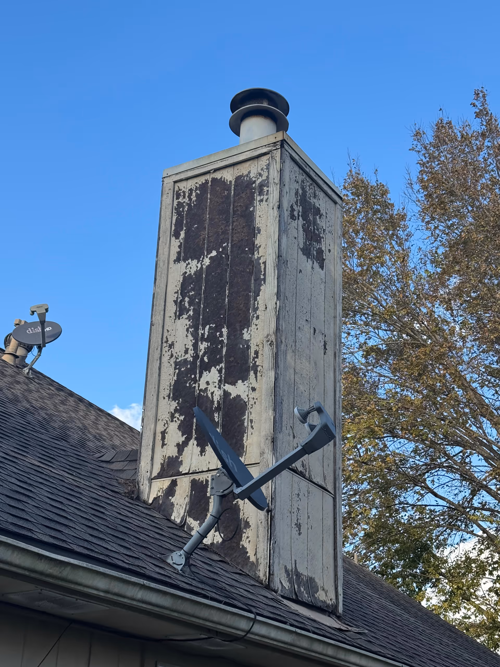 Weathered chimney on a sloped roof with a satellite dish against blue sky