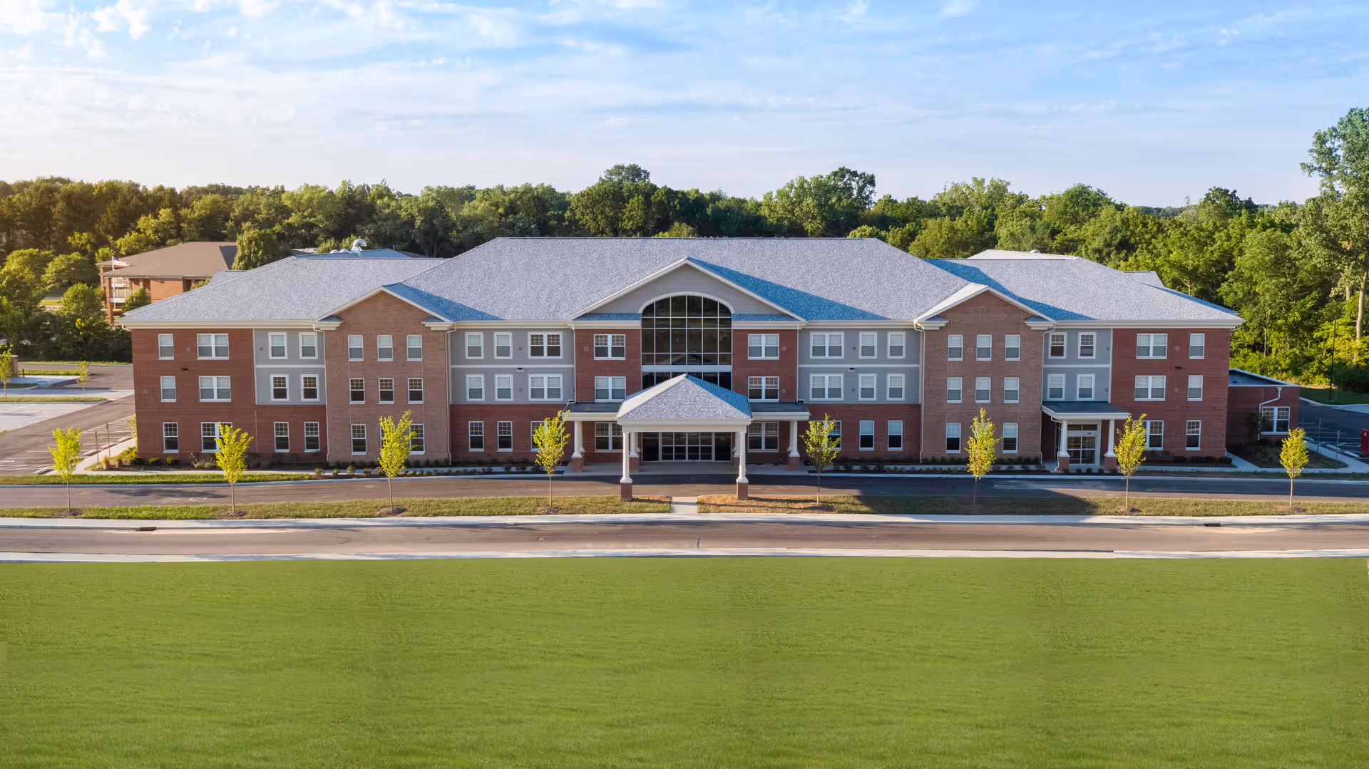 Front exterior view of a large three-story senior living facility building with a covered entrance, multiple windows, and a well-maintained lawn in front. The building is surrounded by trees and has a clear blue sky above.