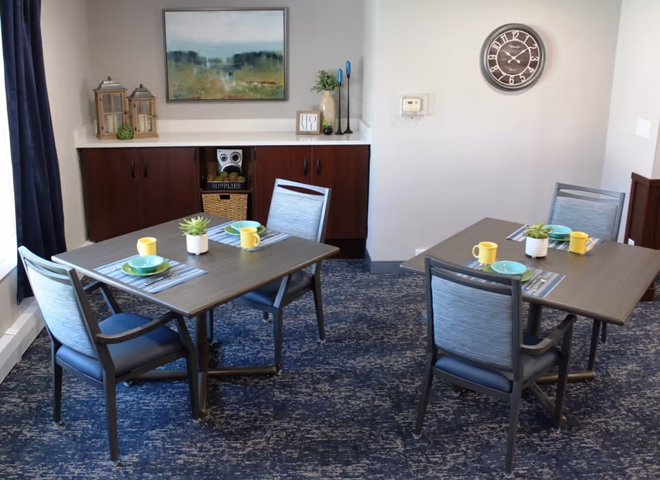 Small communal dining room with two set tables, chairs, decorative centerpieces and a cabinet against the wall.