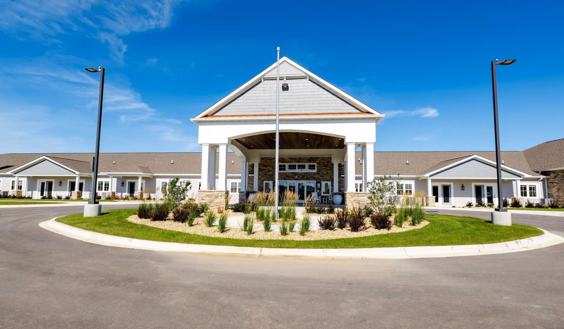 Front entrance of a single-story senior living building with a covered porte-cochere, landscaped circular driveway, and clear blue sky.