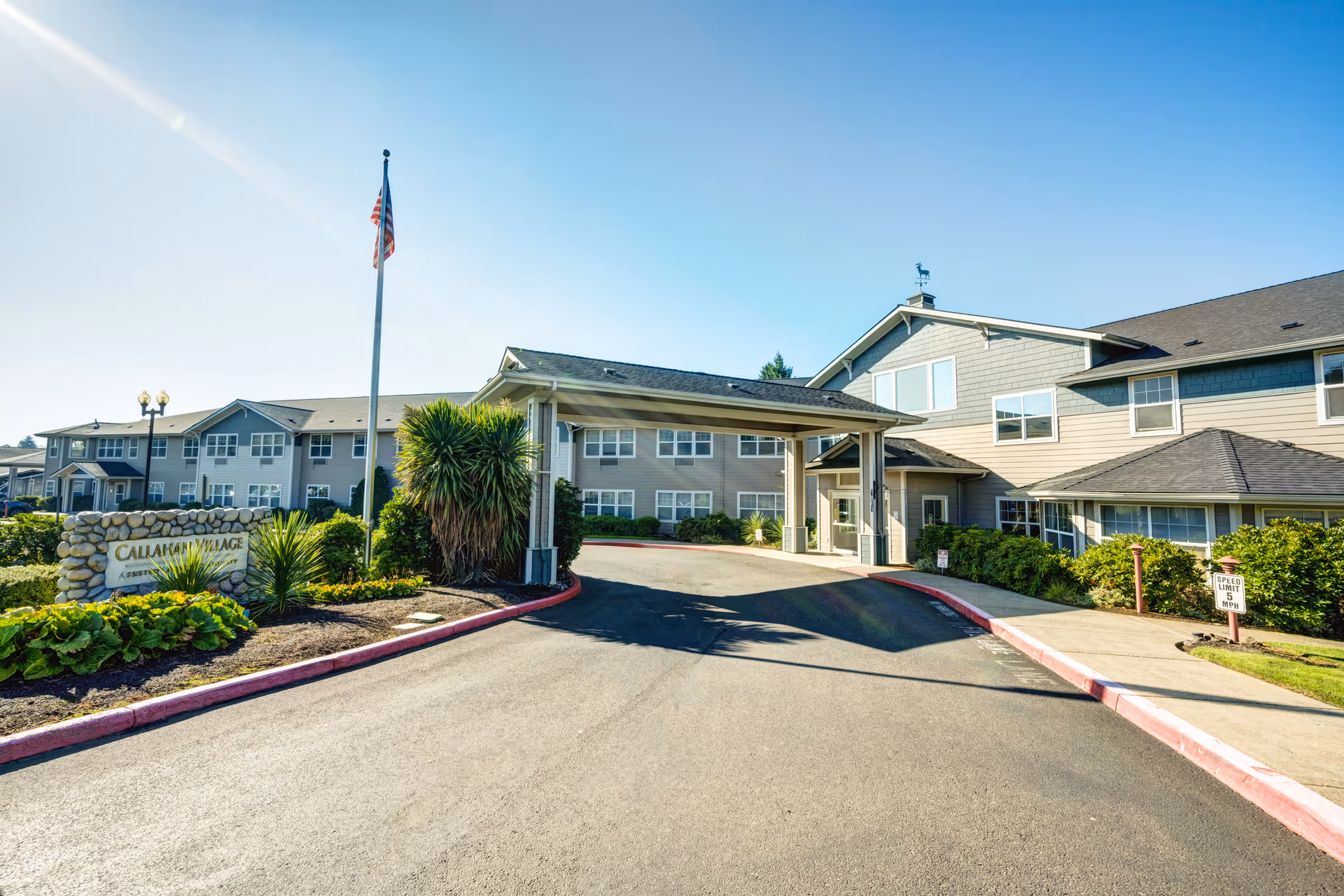 Exterior view of Callahan Village Retirement and Assisted Living facility showing a large driveway leading to a covered entrance. The building is two stories with multiple windows, surrounded by landscaped greenery and an American flag on a flagpole near the entrance.