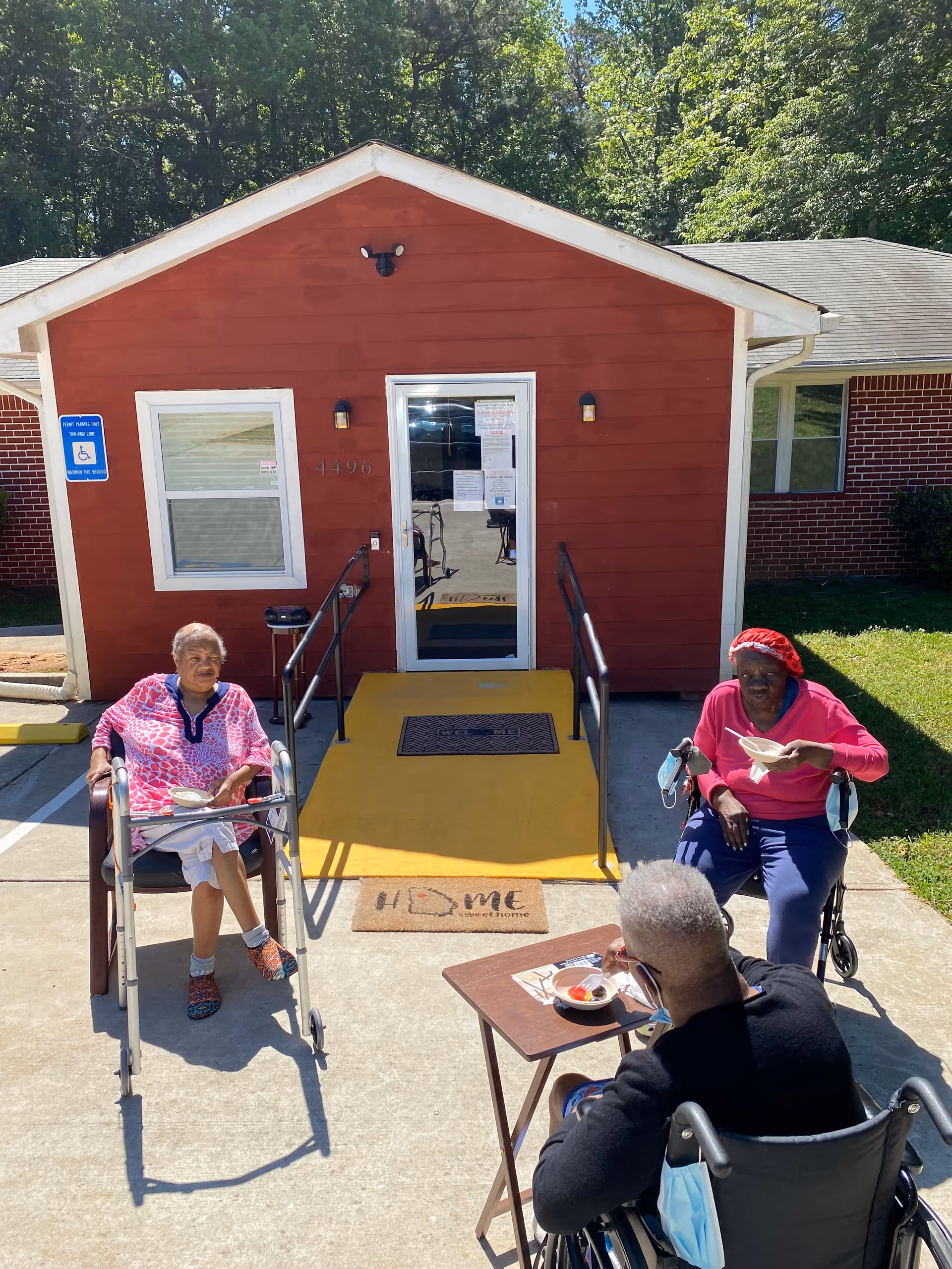 Three seniors in wheelchairs sit outside a red facility entrance, eating at a small table.