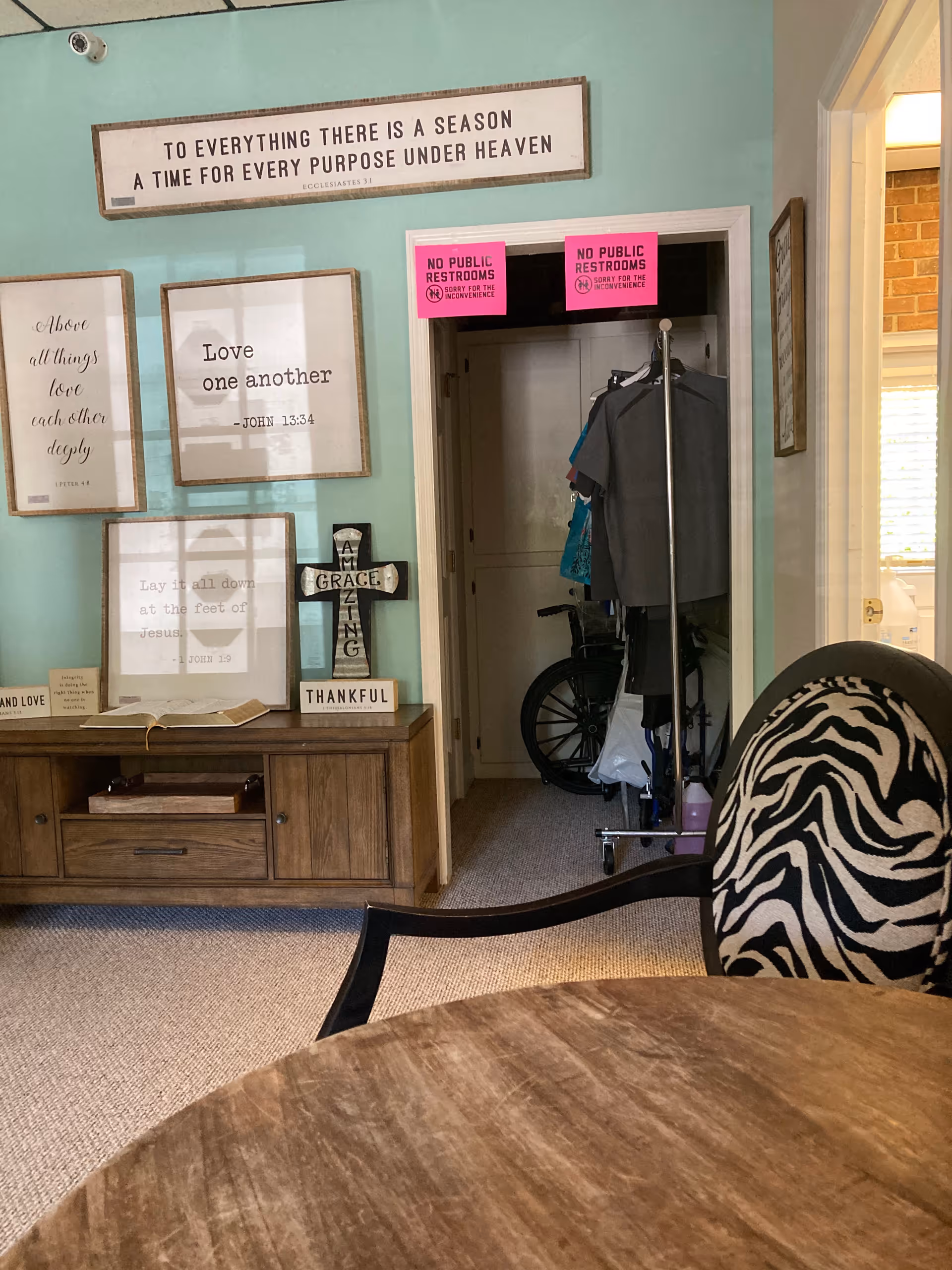 Interior common area shows wall art, a round wooden table, zebra-patterned chair, coats, and a wheelchair.