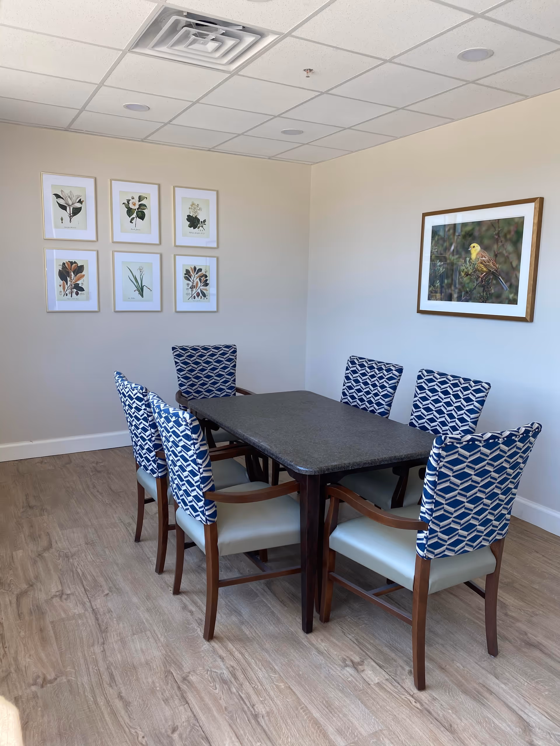 Small dining/meeting room with a rectangular table surrounded by six blue-patterned chairs, botanical and bird artwork on the walls, and wood-look flooring.