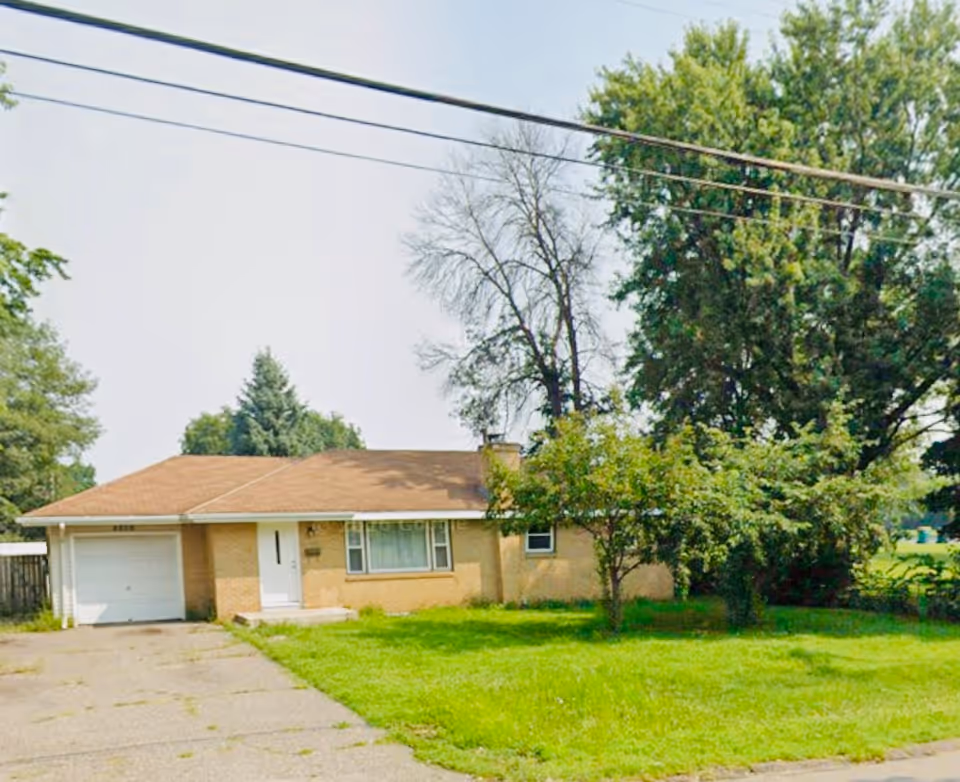 Single-story beige house with attached garage, green lawn, and surrounding trees.