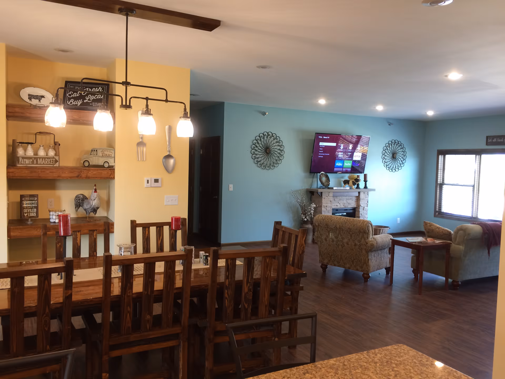 Open-plan living and dining area with a wooden dining table and chairs in the foreground, pendant lights, and a living room with a TV mounted above a fireplace against a blue accent wall.