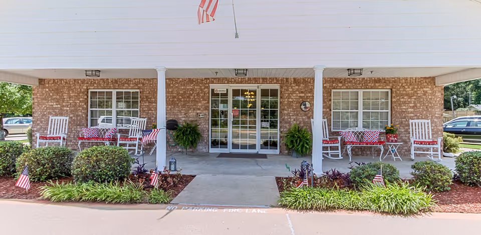 Brick-front covered entrance with double glass doors, rocking chairs, potted plants, and patriotic decorations.