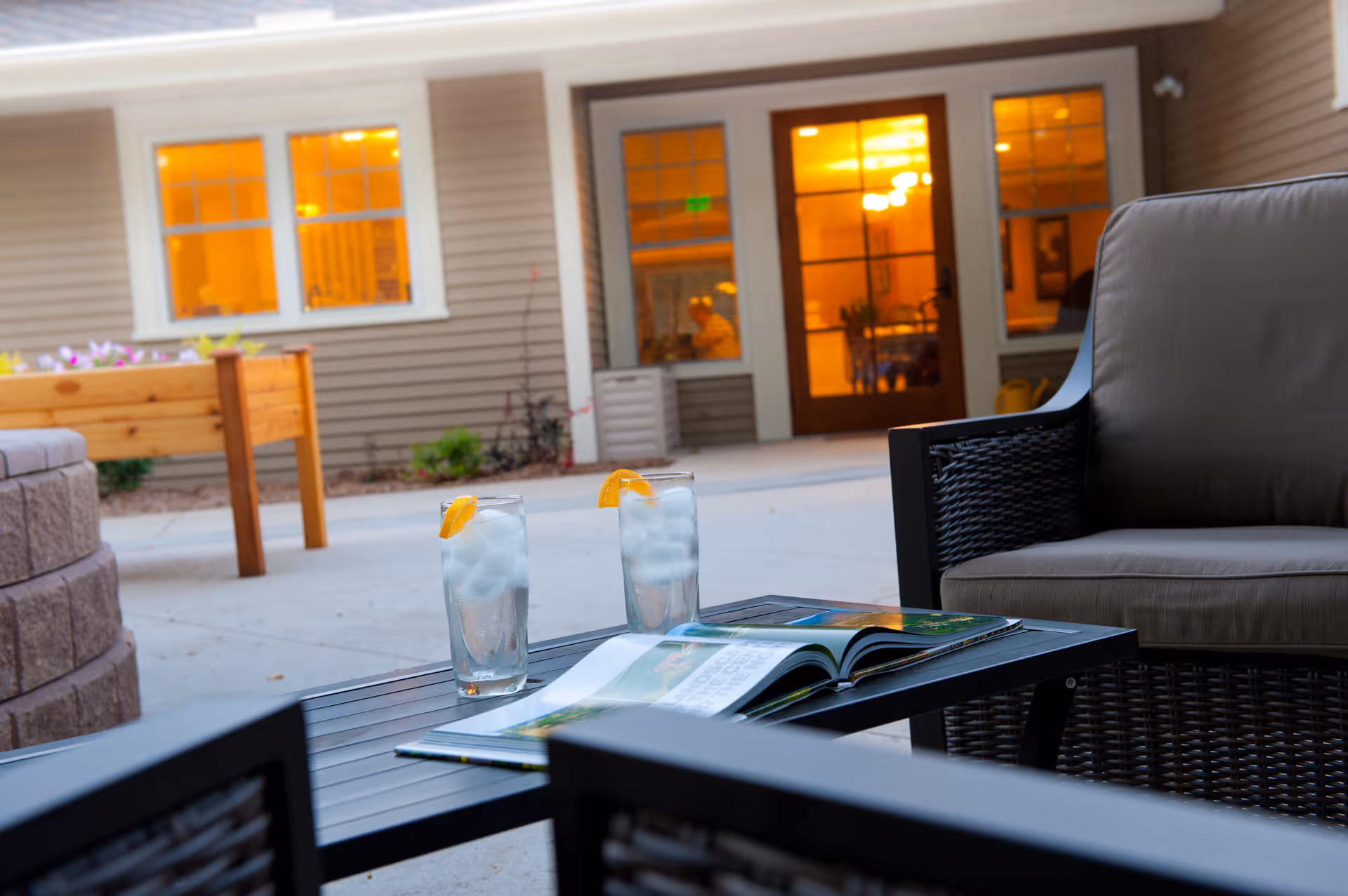 Outdoor patio with two drinks on a table and wicker seating near a lit building.