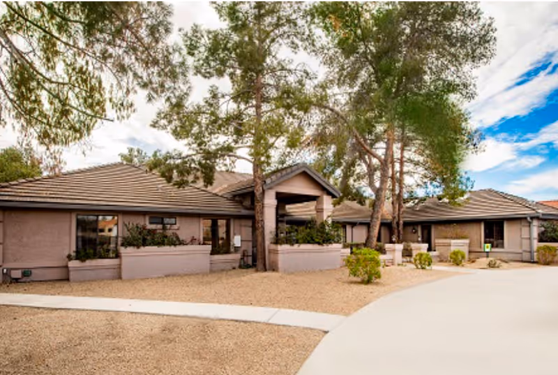 Single-story senior living facility building with a covered entrance, desert landscaping and trees under a partly cloudy sky.