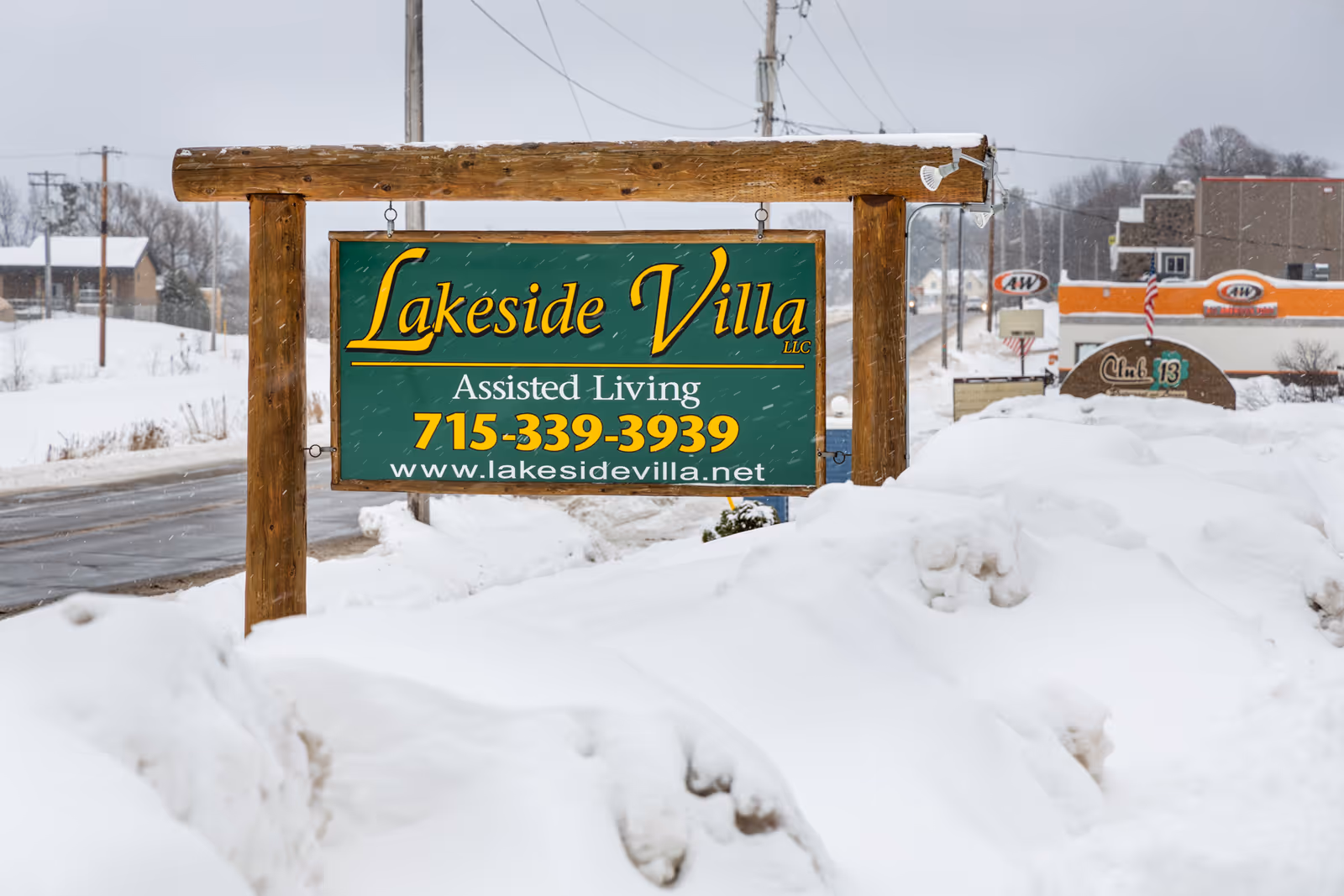 A wooden sign for Lakeside Villa Assisted Living is displayed outdoors in a snowy landscape. The sign includes the phone number 715-339-3939 and the website www.lakesidevilla.net. Snow covers the ground and nearby areas, with a road and some buildings visible in the background.