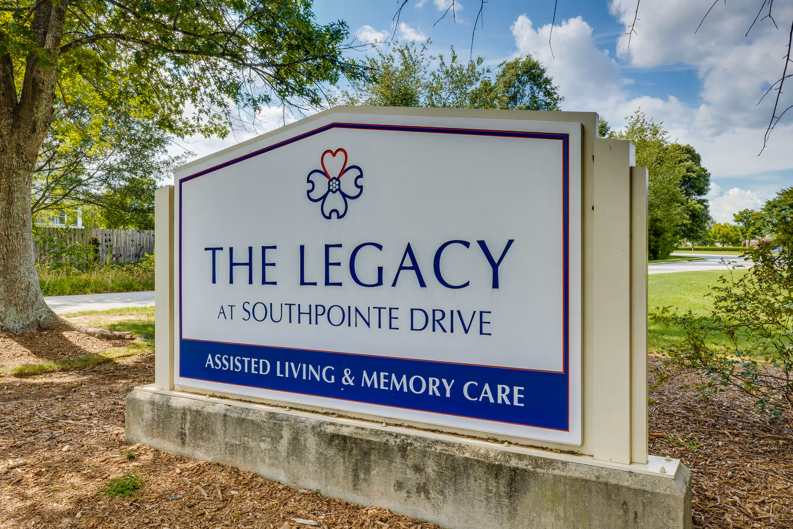 Outdoor entrance sign that reads 'The Legacy at Southpointe Drive - Assisted Living & Memory Care' with trees and sky in the background.