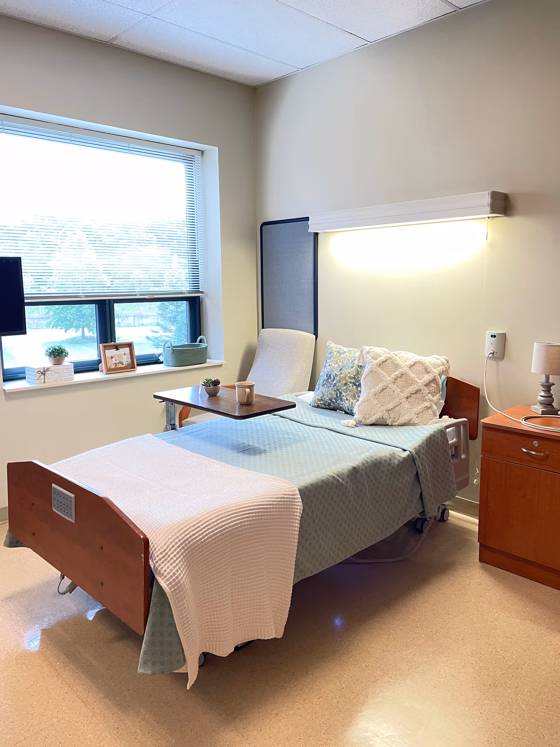 A neatly made hospital-style bed with light blue bedding and decorative pillows in a well-lit room. There is a wooden bedside table with a lamp and a call button on the wall. A window with blinds lets in natural light, and a small table with a cup and plant is positioned over the bed.