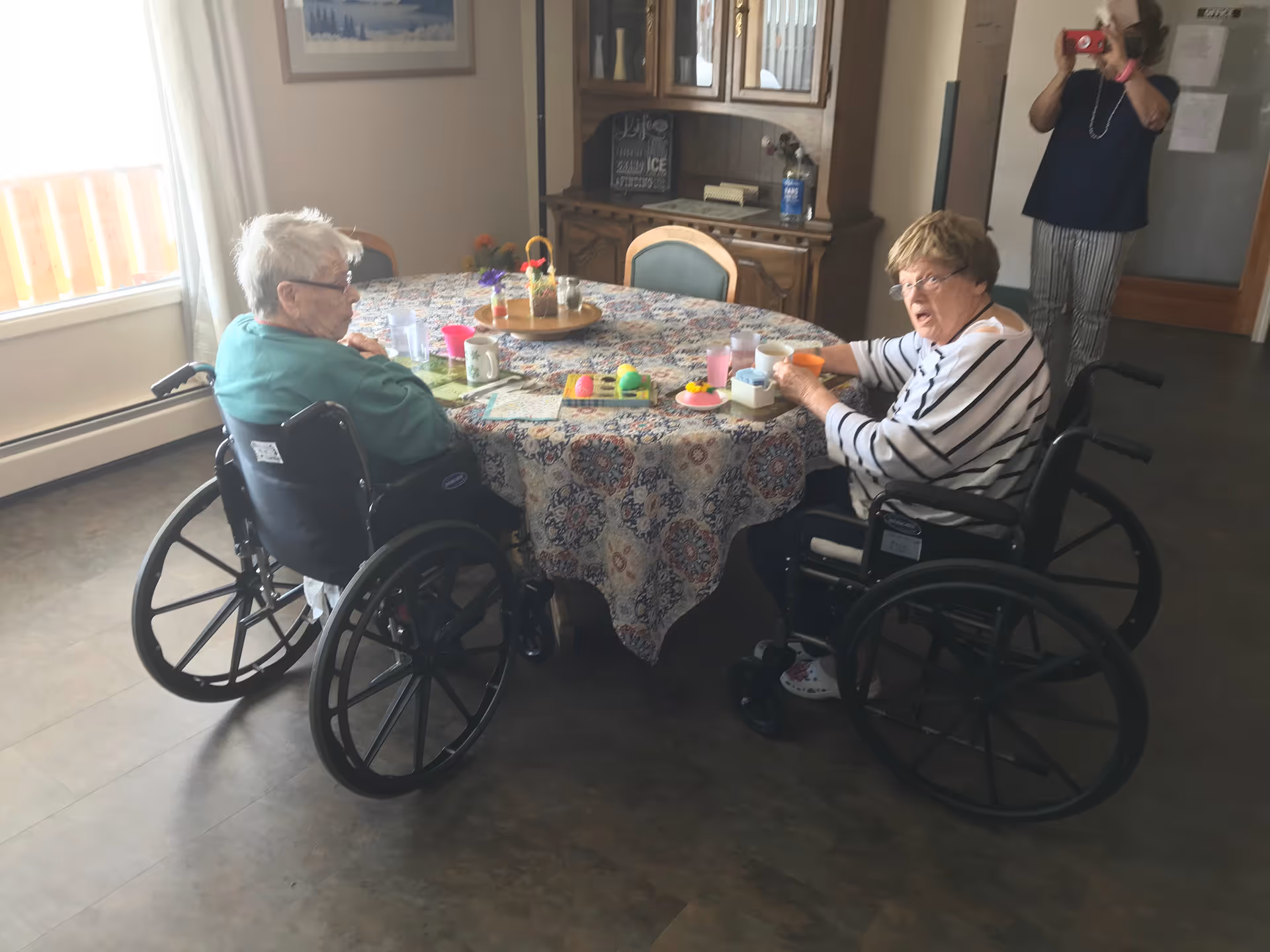 Two elderly women in wheelchairs sitting at a round table covered with a patterned tablecloth, engaging in an activity with colorful objects. A woman in the background is taking a photo with a smartphone. The room has a window with curtains, a wooden cabinet, and a framed picture on the wall.