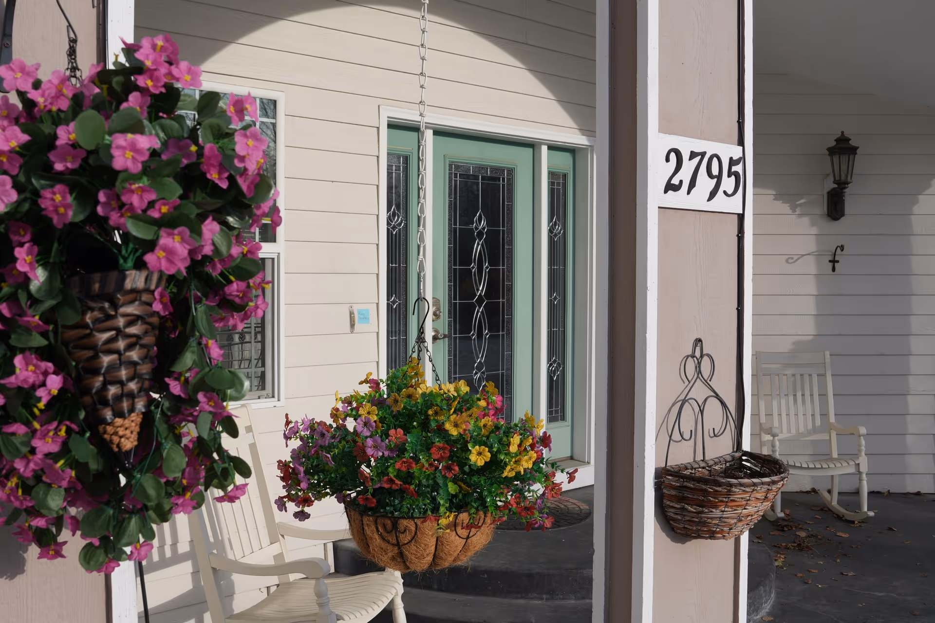 Front porch of a light-colored house with hanging baskets and rocking chairs.