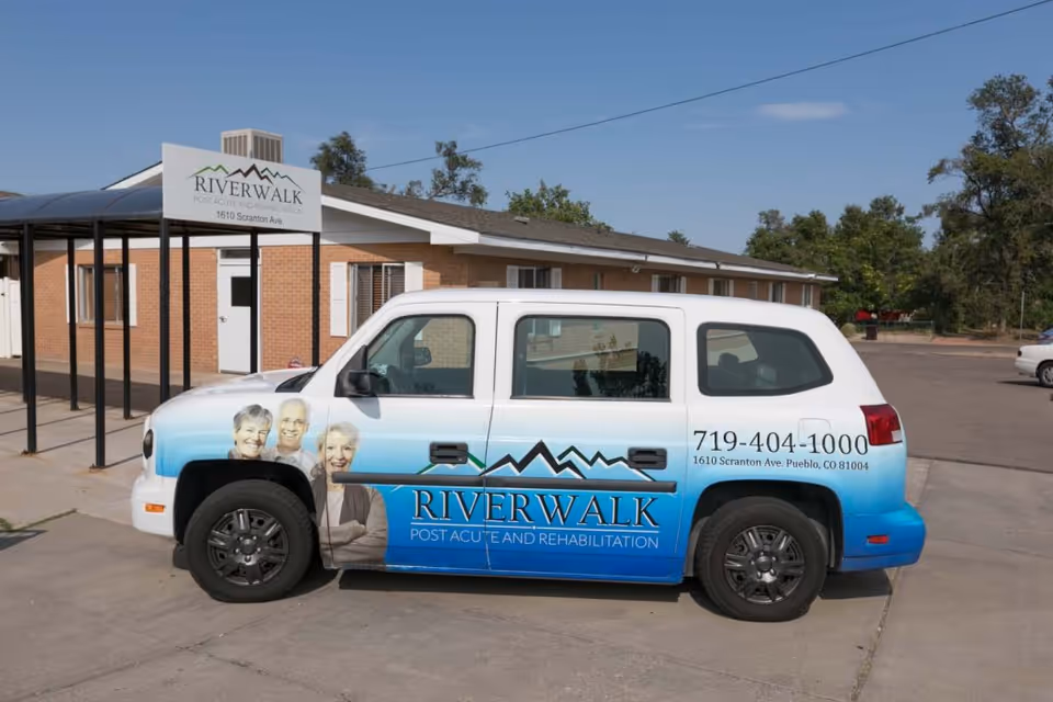A white and blue vehicle parked outside a brick building with a covered walkway. The vehicle has images of three elderly people and text that reads 'Riverwalk Post Acute and Rehabilitation' along with a phone number and address. A sign with the same facility name and address is visible near the building entrance.