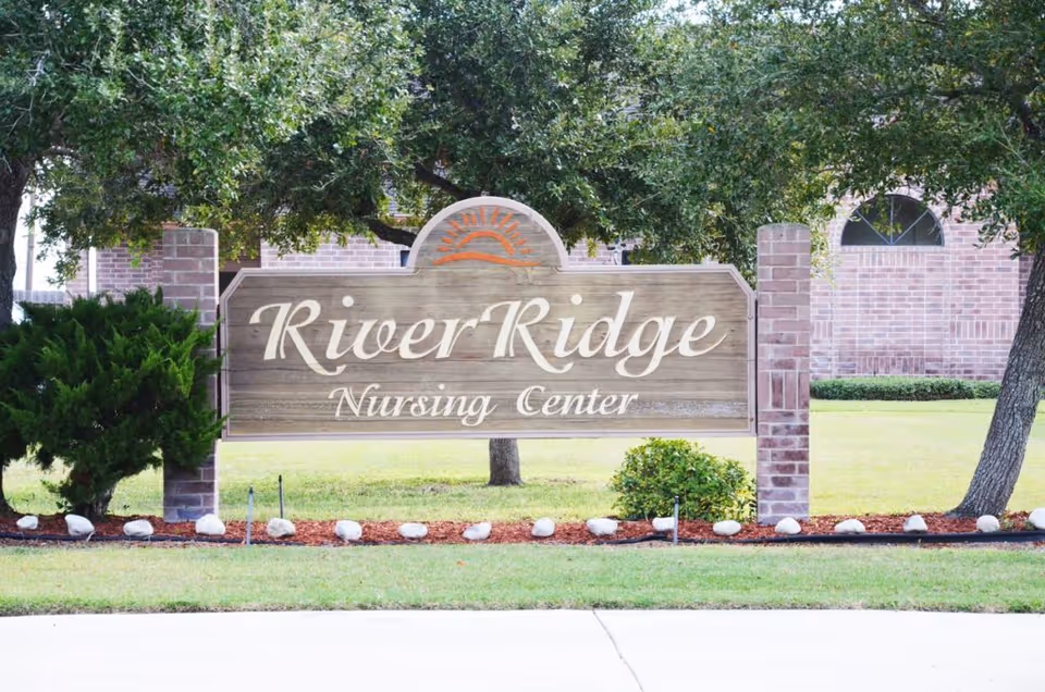Wooden River Ridge Nursing Center sign in front of brick building with trees.