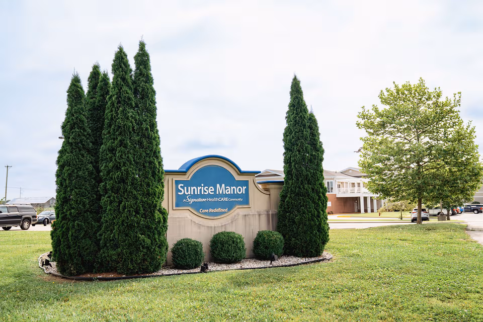 Exterior view of Sunrise Manor Nursing Home sign surrounded by trimmed trees and lawn.