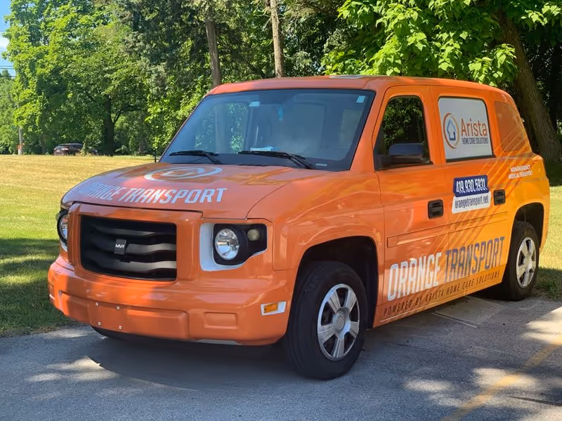 Orange transport van branded Arista Home Care Solutions parked on a grassy, tree-lined area.