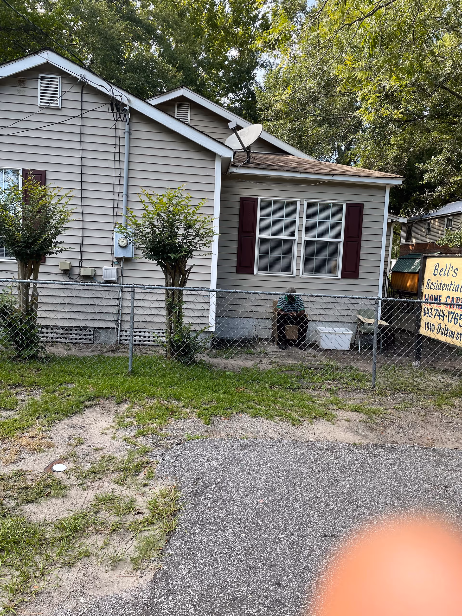 Exterior view of a single-story residential building with beige siding and maroon shutters on the windows. There is a chain-link fence in front of the building with two small trimmed trees. A person is sitting on a chair near the window, and a yellow sign on the right side reads 'Bell's Residential Home Care' with a phone number and address. The area has some grass and a paved driveway or road in the foreground.
