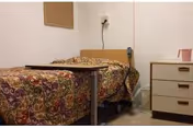 A bedroom in a nursing home with floral quilt on bed, bedside table, and dresser.