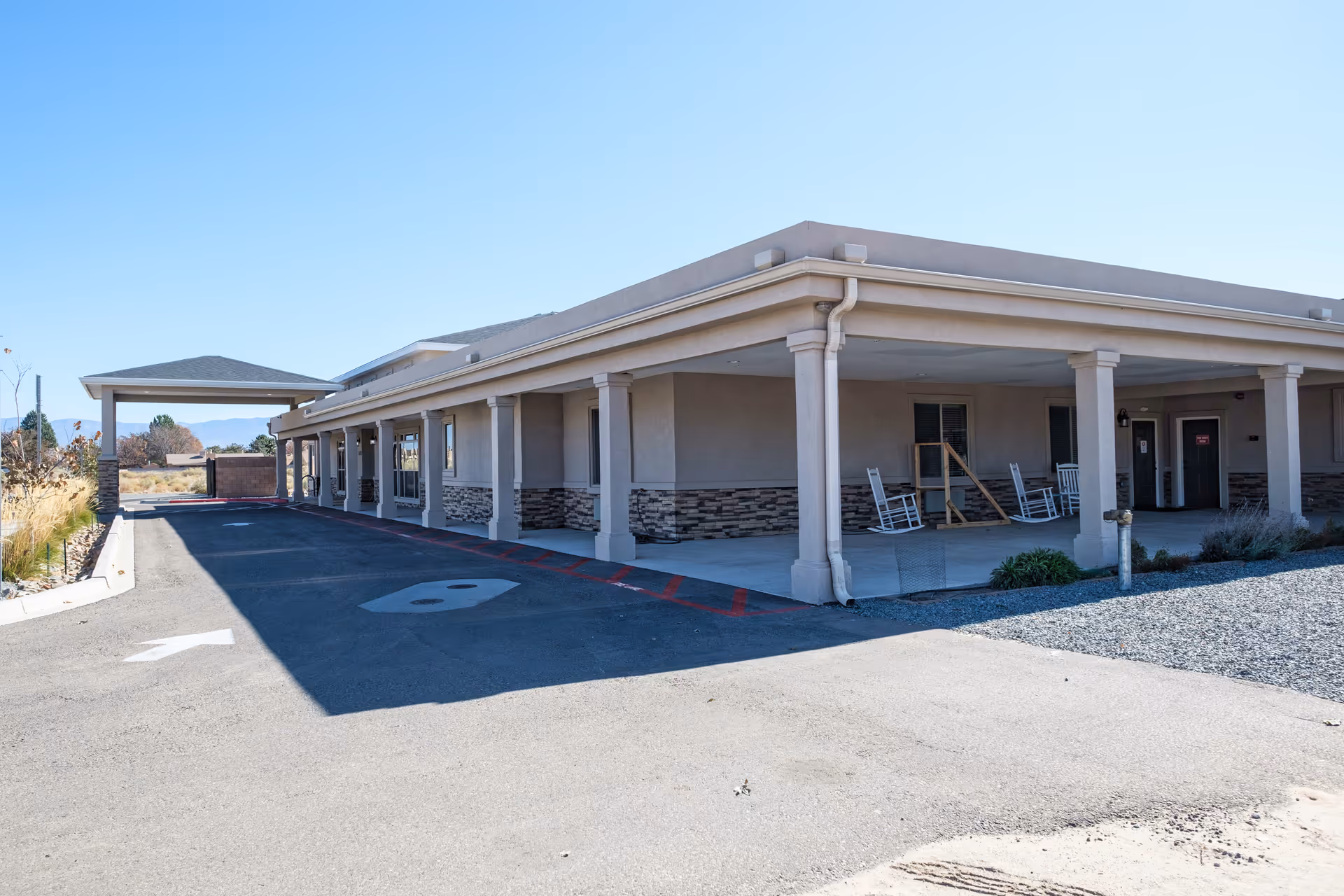 Exterior view of a single-story senior living facility with a covered porch and chairs.