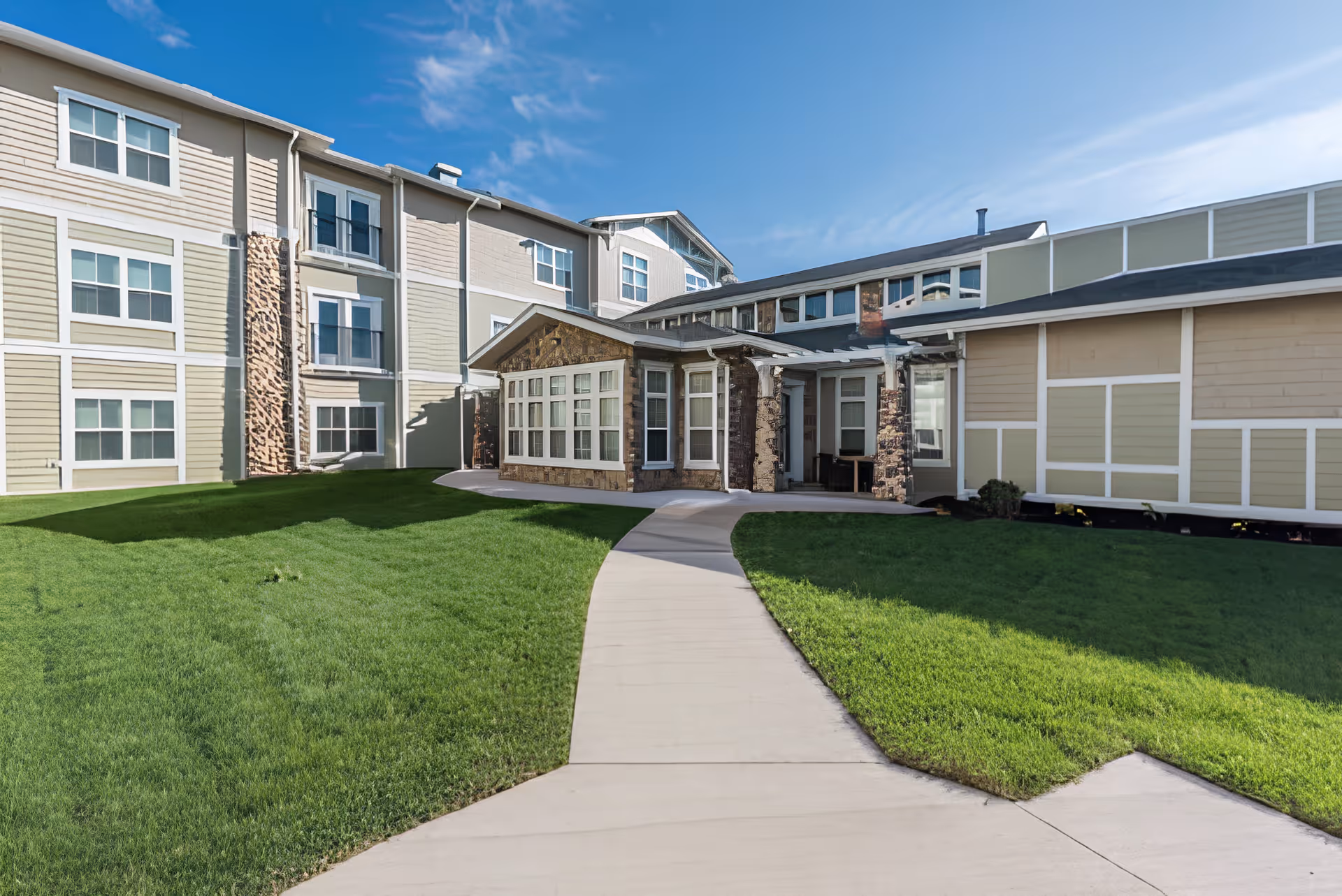 Exterior view of a three-story senior living building with a paved walkway leading to the main entrance and green lawns on either side.