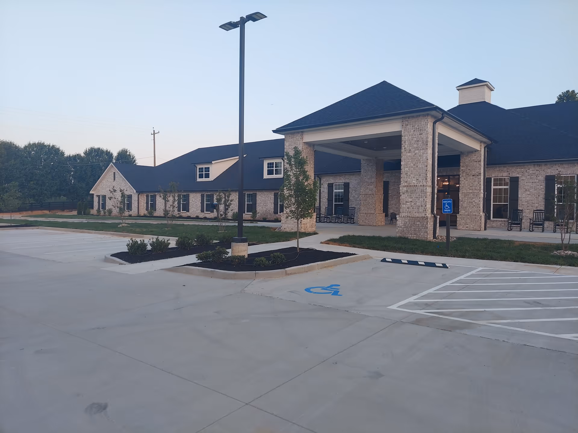 Exterior view of The Pearl at Fairview facility showing a large brick building with a covered entrance, several windows, rocking chairs on the porch, a parking lot with handicap parking spaces, and a tall street lamp. Trees and landscaping surround the building.