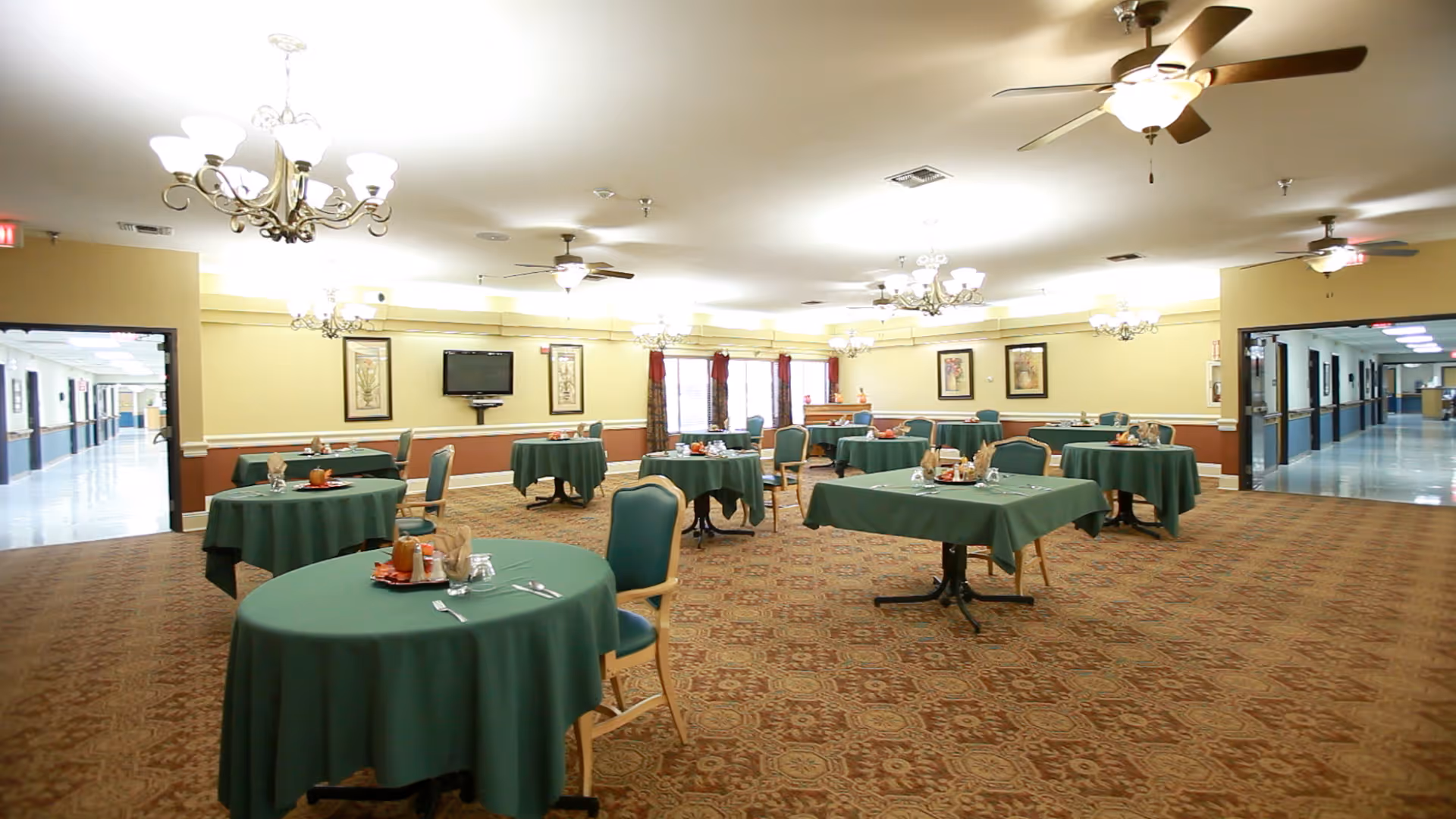 A spacious dining room with multiple round tables covered with green tablecloths, each set with plates, glasses, and utensils. The room has beige walls with framed artwork, carpeted floors, ceiling fans, and chandeliers providing lighting. Two open doorways lead to hallways on either side of the room.