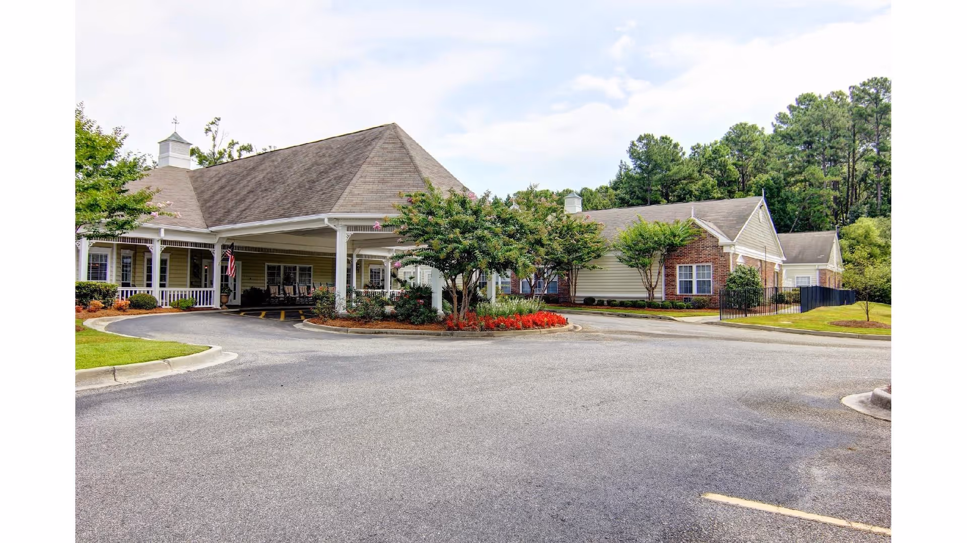 Exterior view of The Crossings of Georgetown senior living facility showing a large building with a covered entrance, surrounded by landscaped greenery and trees under a partly cloudy sky.